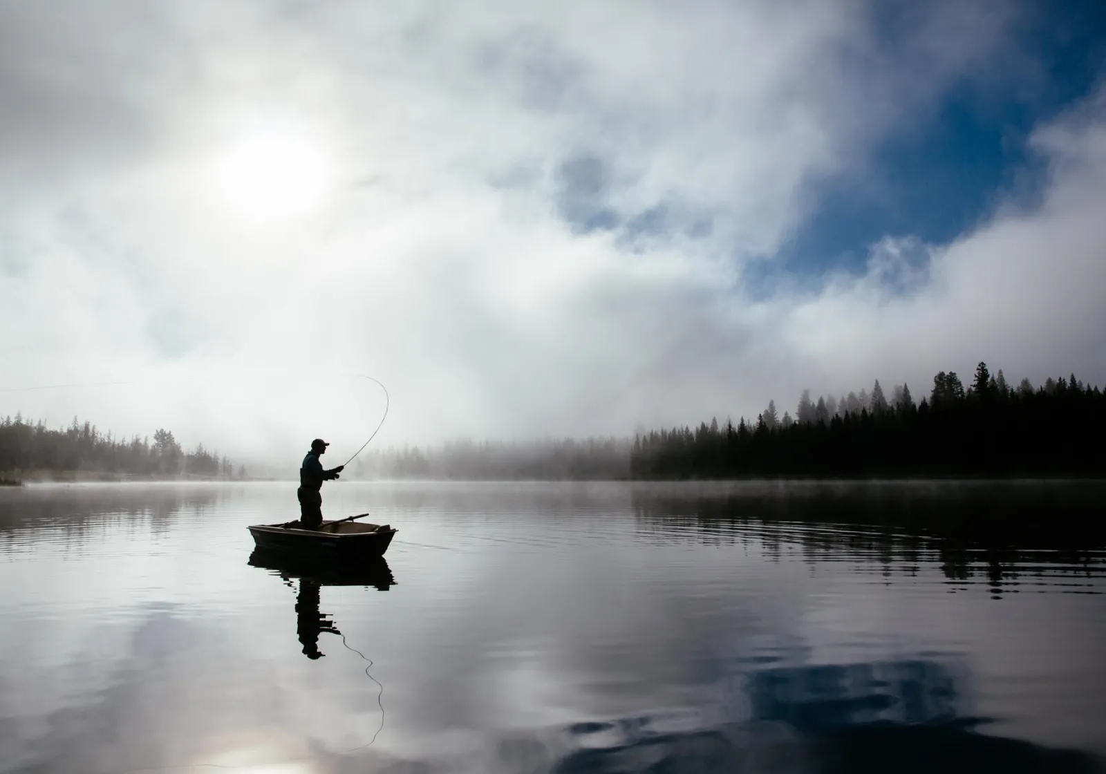 A fisherman stands in a boat on a serene wilderness lake while fly-casting for rainbow trout